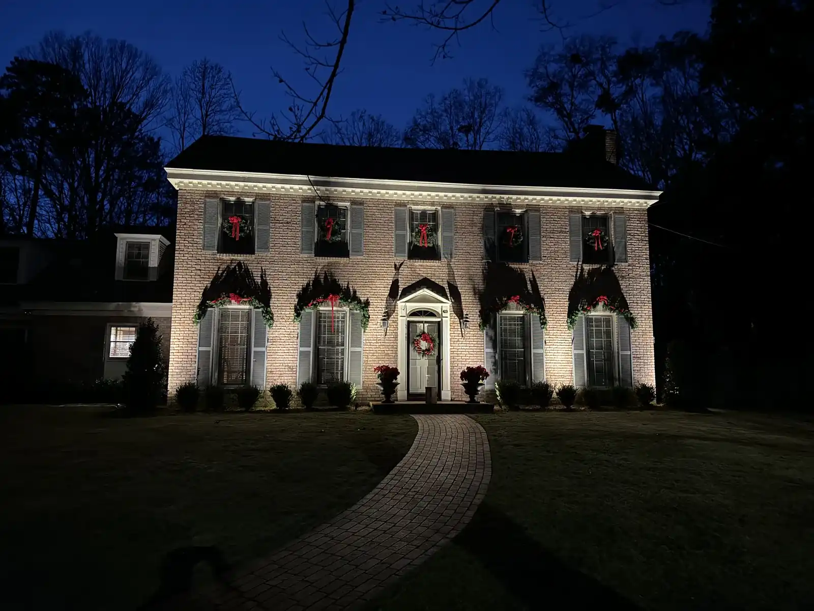 Two-story brick colonial home with uplighting on the front facade and a curved walkway at night
