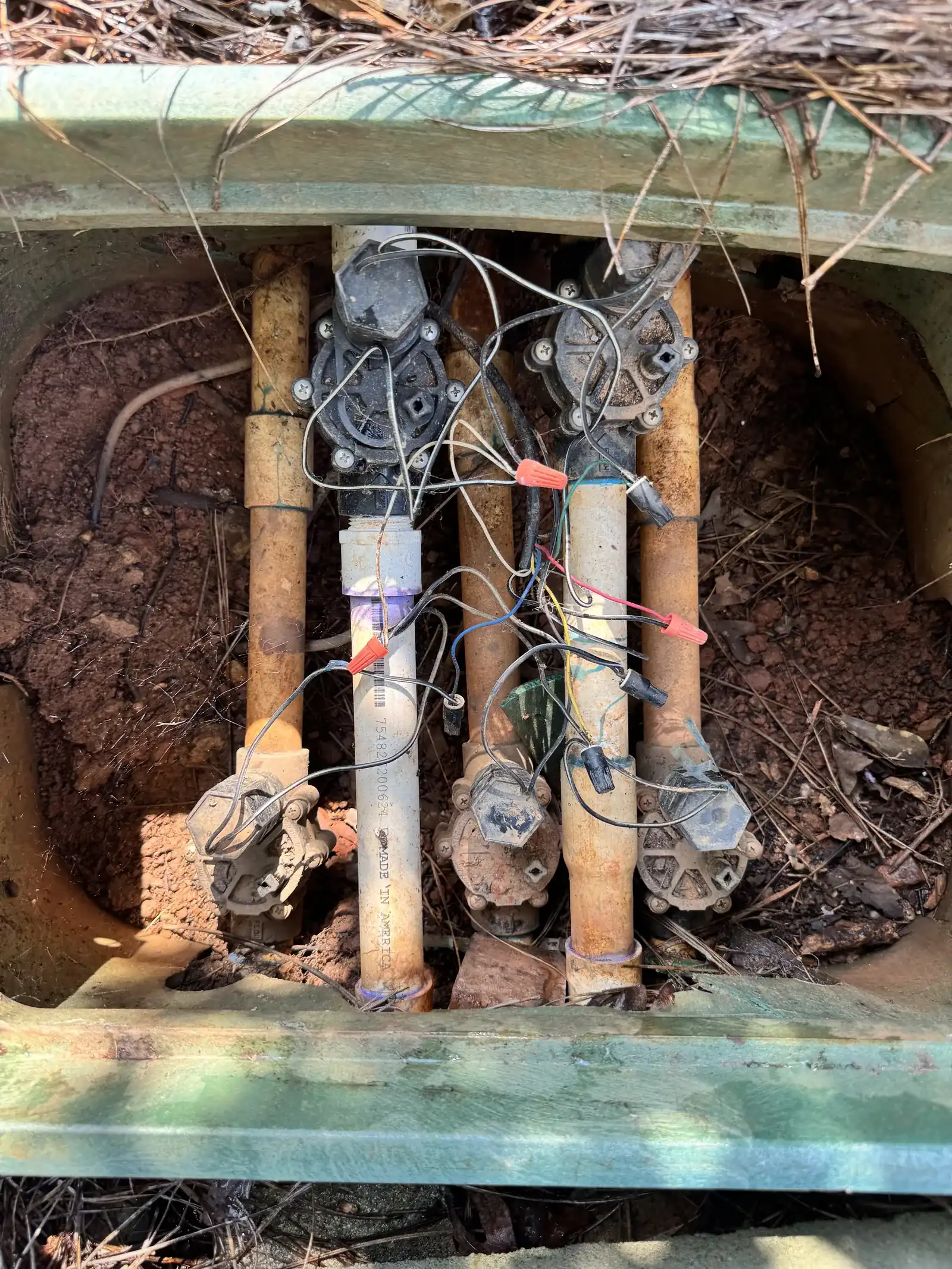 Open valve box with a row of old irrigation valves and brass and PVC piping before repair